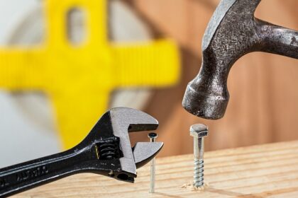 Hammer knocking in a screw into a wooden board beside a spanner extracting a nail from the same wooden board